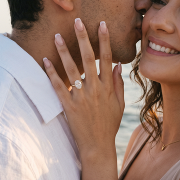A close-up lifestyle shot of a couple embracing on a beach at sunset, with the woman wearing a silver beaded bracelet and a silver engagement ring with a round clear gemstone.