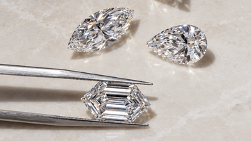 Five loose diamond cuts in different shapes arranged on a light stone surface, with one baguette-cut diamond held in silver tweezers. No person or real environment is present beyond a simple styled surface, so this fits Product/Packaging rather than Lifestyle.